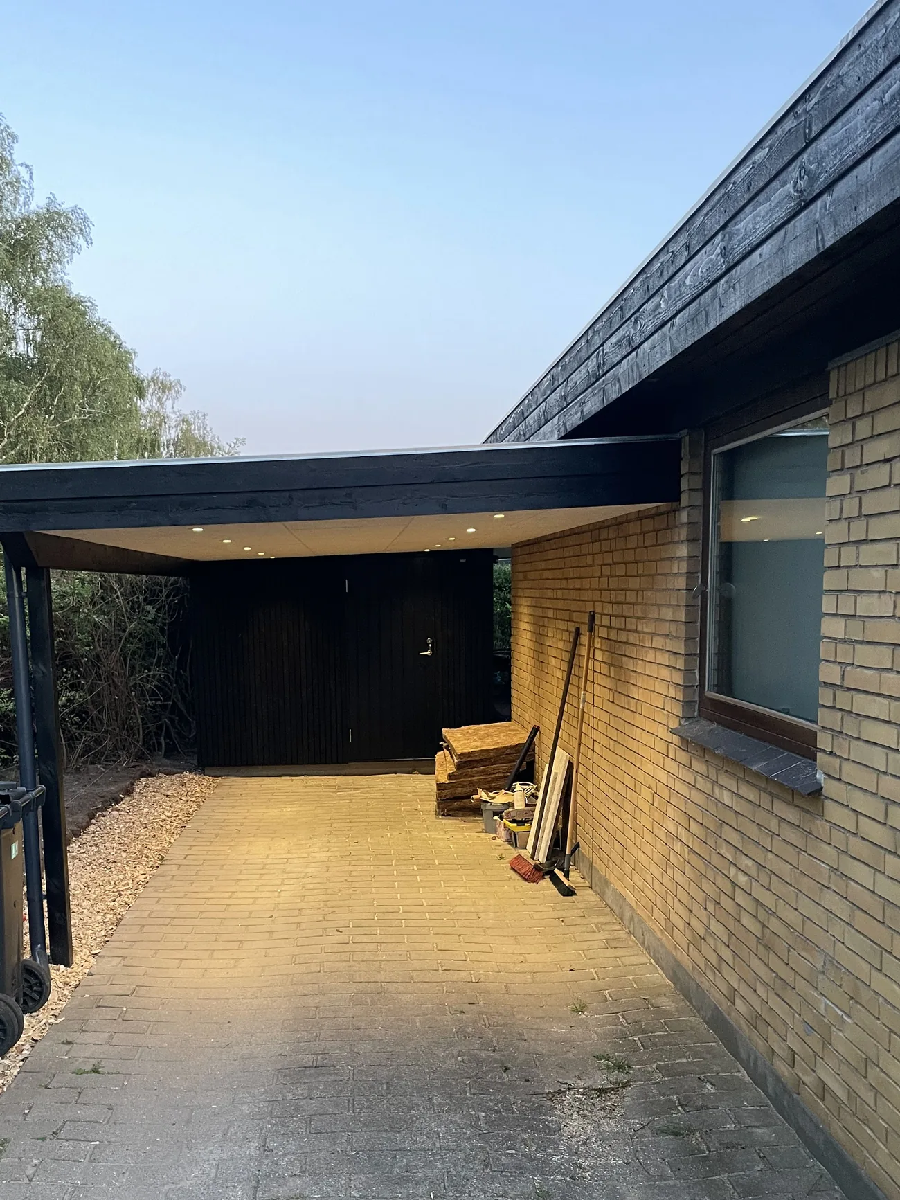 Sortmalet carport med fladt tag, indbyggede spots og skurdør for enden, bygget op ad husets gavl